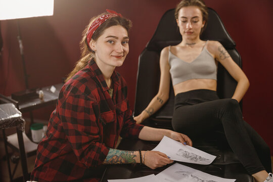 Smiling tattoo artist looking at camera while holding paper with picture in hands