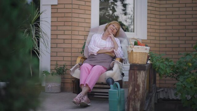 Wide Shot Senior Woman Dreaming Sitting In Rocking Chair On Porch Outdoors. Portrait Of Positive Caucasian Female Retiree Enjoying Leisure On Front Yard Backyard Looking Away Smiling. Slow Motion