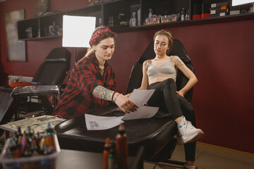 Focused woman tattoo master holding sketches for tattoo on paper and showing to female client sitting next to her