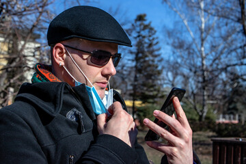 The young man holding down his face mask to make a video call on his smartphone. Outdoors. Coronavirus protection measures.