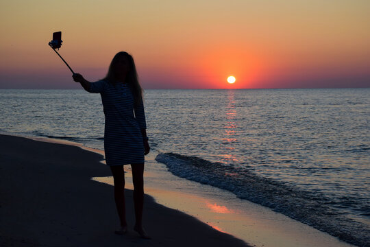 At sunset on the beach, a silhouette of a woman takes selfie pictures on a mobile phone