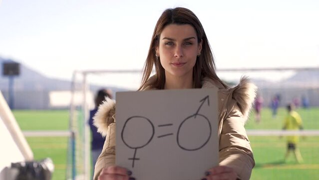 Close-up shot of a young woman holding a paper with a symbol of gender equality drawn on it, in front of a soccer field in slow motion