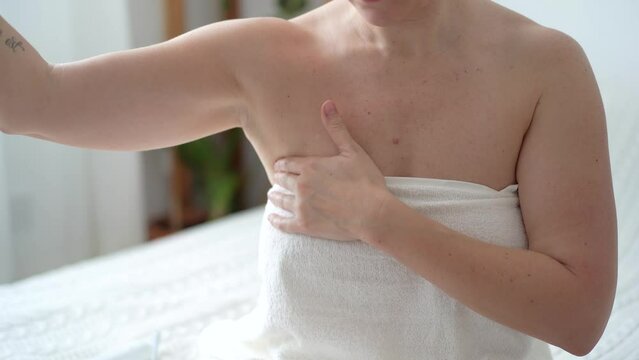 Young woman examines her breasts and lymph nodes with her hands while sitting at home in the bedroom after a bath. Prevention of diseases of the female breast