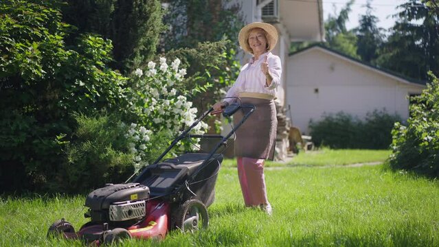 Wide shot portrait of positive senior woman gesturing thumb up smiling standing with lawn mower. Confident Caucasian retiree posing in sunshine on green backyard lawn. Lifestyle and confidence