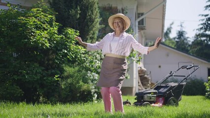 Wide shot cheerful senior woman dancing in slow motion on sunny summer lawn on backyard outdoors. Portrait of joyful happy Caucasian retiree having fun in sunlight on green meadow. Retirement and joy