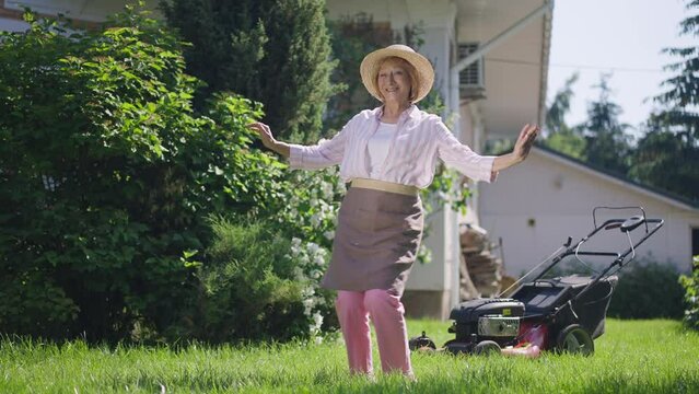 Wide shot cheerful senior woman dancing in slow motion on sunny summer lawn on backyard outdoors. Portrait of joyful happy Caucasian retiree having fun in sunlight on green meadow. Retirement and joy