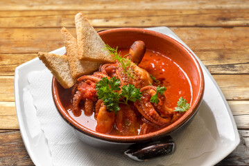 Octopus in tomato soup with toast and parsley in earthenware on wooden table