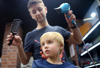 Preschooler boy getting haircut in barbershop. Children hairdresser with professional tools - comb and hairdryer. Cutting hair for kids