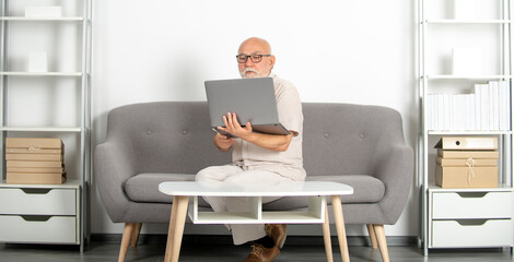 Mature consultant, senior man in office. Old man sits at desk on sofa with laptop, portrait. Older senior business man, grey-haired man wearing glasses looking at laptop.