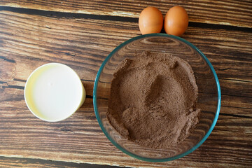 a glass bowl with rice flour, sugar and cocoa stands on a wooden table next to two eggs and a glass of coconut milk. top view. recipe and preparation of vegan cupcake