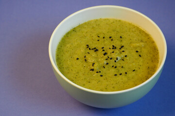 on a lilac background, a green plate with green broccoli soup, sprinkled with black sesame seeds. vegan soup without gluten and lactose. healthy food