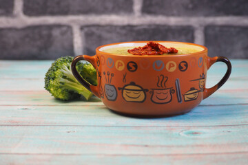 on a blue wooden table there is a brown plate with green broccoli puree soup with dried tomatoes on top next to sprigs of fresh broccoli. vegan soup without gluten and lactose. healthy food