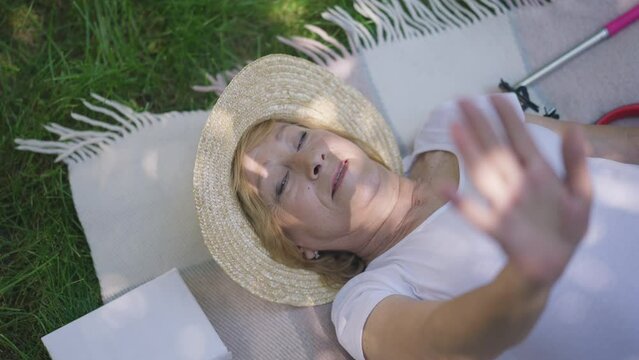 Top view happy senior woman in straw hat lying on blanket in park looking at sunshine through fingers. Portrait of relaxed female Caucasian retiree enjoying spring summer leisure outdoors