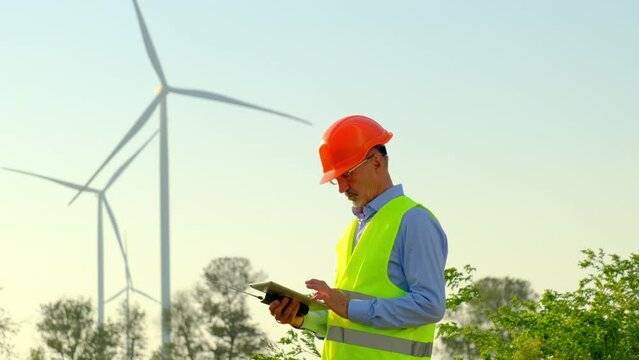 Wind turbines produce alternative energy. Engineer in orange hardhat verifying data via tablet at construction site of rural substation
