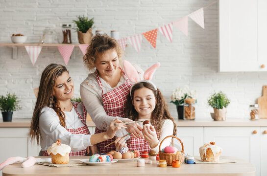 A happy mom with her adult daughters paints Easter eggs at home in the kitchen.