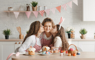 A happy mom hugs her daughters at the kitchen table while preparing for the Easter holiday. Family happiness and joy.