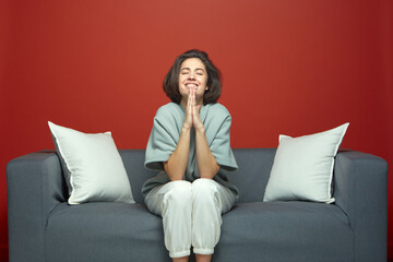 Young woman beg for dream, make praying gesture. Female imploring appealing, sitting on couch on...