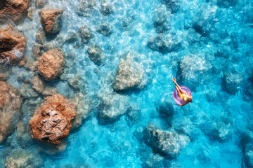 Aerial view of a young woman swimming with pink swim ring in blue sea at sunset in summer. Tropical landscape with girl, clear water, stones, sandy beach. Top view. Vacation in Lefkada island, Greece