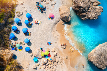 Aerial view of blue sea, sandy beach with umbrellas, stones and rocks in water at sunset in summer. Lefkada island, Greece. Tropical landscape with sea coast, swimming people, blue water. Top view