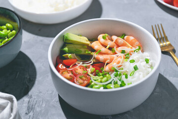 Poke bowl with red shrimps, rice, tomatoes, avocado and edamame beans in the white bowl on the gray background. Closeup.