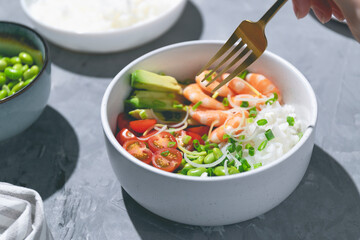 Poke bowl with red shrimps, rice, tomatoes and edamame beans in the white bowl on the gray background. Closeup.
