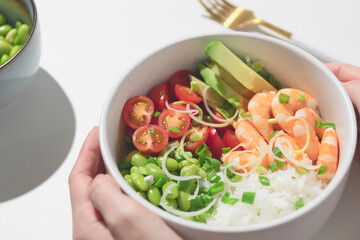 Woman holding poke bowl with red shrimps, rice, tomatoes and edamame beans in the bowl.