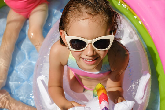 A wet child in a swimsuit and sunglasses is sitting in an inflatable swimming pool in the hot scorching sun. The girl looks directly into the camera. Summer entertainment on leisure