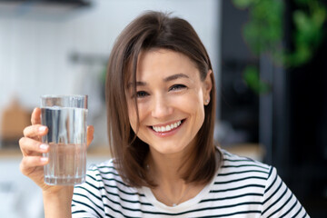 Happy young woman holding glass of water, looks at the camera and smiles friendly. Healthy lifestyle concept