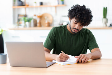 Indian young man using laptop for watching webinar, studying online and takes notes in a notebook, male student is studying online, e-learning concept