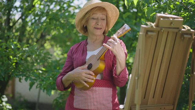 Medium shot happy Caucasian retiree playing ukulele standing in sunshine at easel outdoors in park. Portrait of relaxed talented confident woman enjoying hobbies in garden on sunny summer day