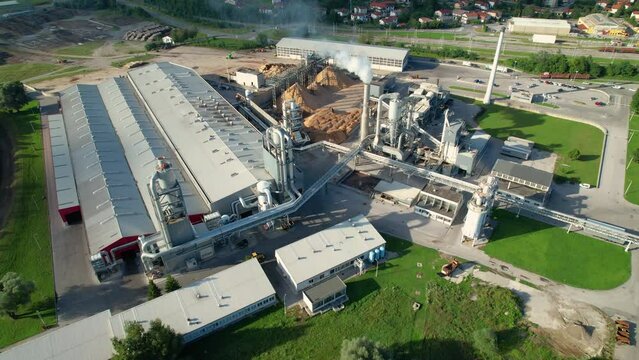 AERIAL: Flying above a large sawmill emitting white smoke from a tall chimney. Drone view of a large logging factory with a tin-roofed processing facility. Warehouse and timber processing factory.