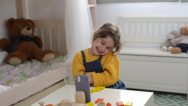 Funny little girl posing on smarthpone video doing dance sitting in room and laughing. Kids using phone app making selfie as a game