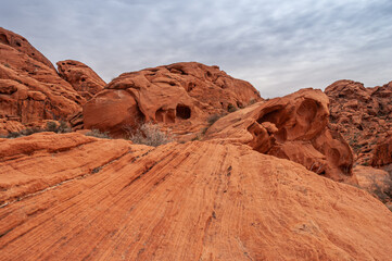 Overton, Nevada, USA - February 25, 2010: Valley of Fire. Landscape of naturally shaped holes and caves in red rock boulder under gray cloudscape. Few bushes on red lined desert floor. 