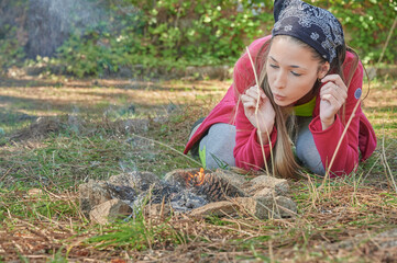 Young college girl wearing mountain clothes blows to burn some pine needles and start a small...