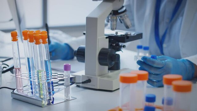 Close up of scientist wearing white coat looking through microscope and making notes whilst holding test tube which he puts on desk to show label omega - shot in slow motion