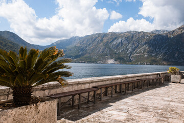 Perast, Montenegro - 15.09.2021: Roman catholic church our lady of the rocks on island islet in boka kotor bay in front of mountains range and blue sky. Unesco world heritage. Palms grows here