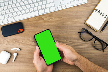 Man holding a smartphone with green screen on the wooden table. Office environment. Chroma Key