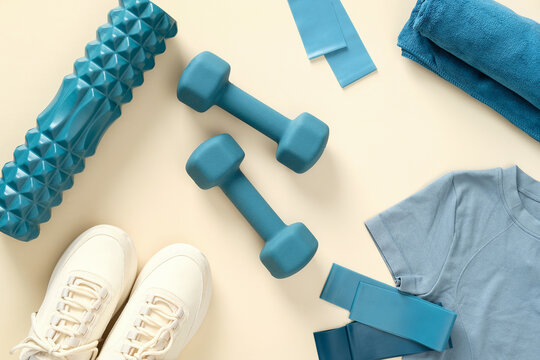 Fitness and workout equipment top view. Flat lay dumbbells, exercise roller, elastic bands, feminine sneakers, t-shirt on beige table.