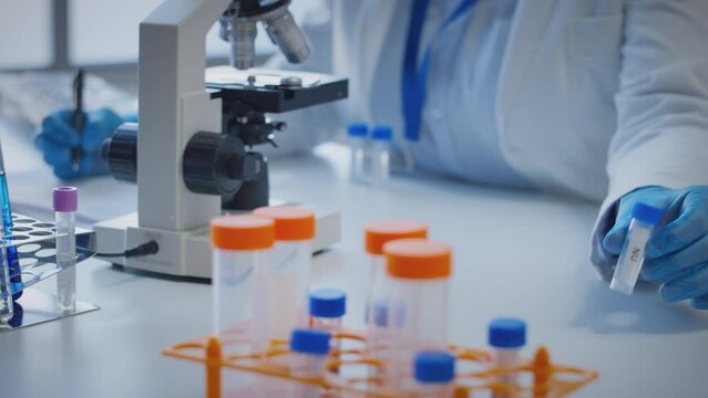 Close up of scientist wearing white coat looking through microscope and making notes whilst holding test tube which he puts on desk to show label nu - shot in slow motion