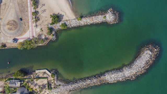 Aerial photos of a wavebreaker, taken from a drone, where you can see the breakwater at 90 degrees, rocks, the greenish sea, tide, plants, trees, buildings
