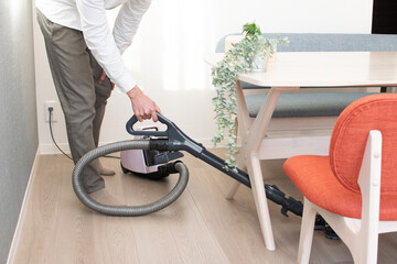 A Japenese young Asian man hoovers the floor using a vacuum cleaner in a house with a desk and two chairs