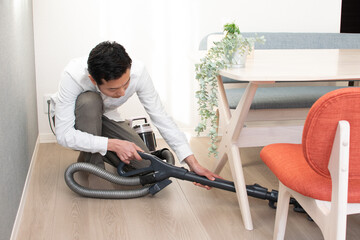 A Japenese young Asian man hoovers the floor using a vacuum cleaner in a house with a desk and two chairs