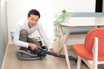A Japenese young Asian man hoovers the floor using a vacuum cleaner in a house with a desk and two chairs