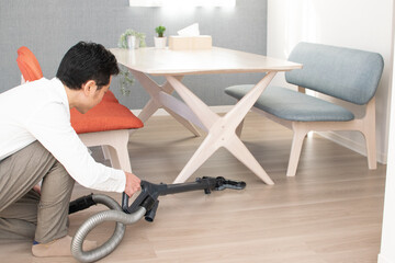 A Japenese young Asian man hoovers the floor using a vacuum cleaner in a house with a desk and two chairs