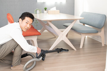 A Japenese young Asian man hoovers the floor using a vacuum cleaner in a house with a desk and two chairs