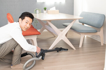 A Japenese young Asian man hoovers the floor using a vacuum cleaner in a house with a desk and two...