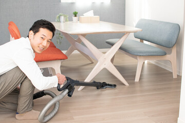A Japenese young Asian man hoovers the floor using a vacuum cleaner in a house with a desk and two chairs