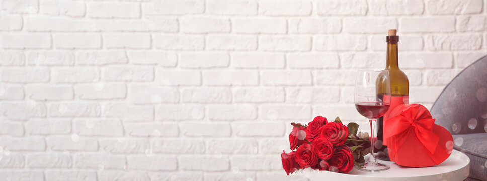 Gift for Valentine's Day with flowers and wine on table against white brick wall with space for text. Valentine's Day celebration