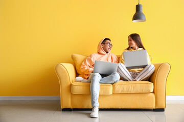 Young couple with laptops sitting on sofa near yellow wall
