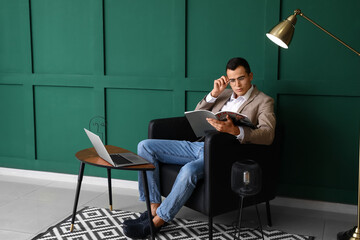 Young man with eyeglasses reading magazine in leather armchair near green wall - obrazy, fototapety, plakaty